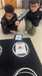 Two male students kneeling near a rug on which they're completing a coding project.